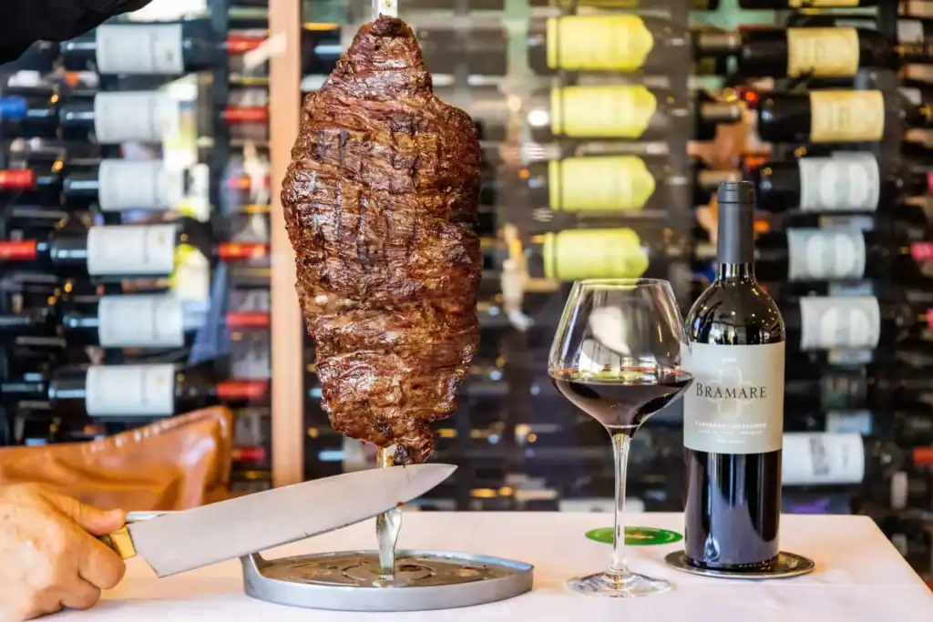 Juicy grilled meat served at Adega Gaucha with wine and wine rack background.