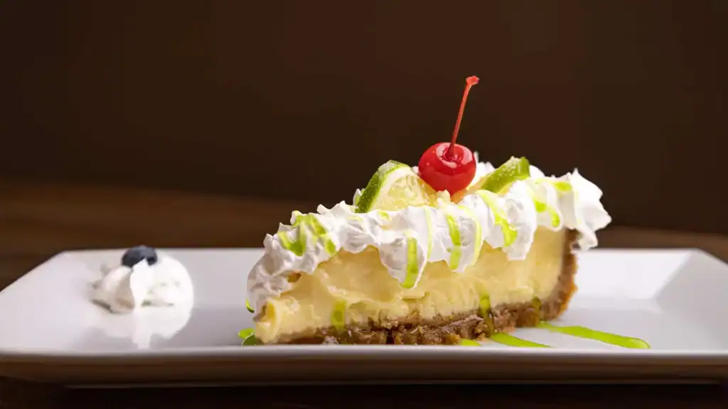 Image description only: A slice of cheesecake topped with lime slices, a cherry, and whipped cream, presented on a white plate, highlighting fresh ingredients and dessert appeal.