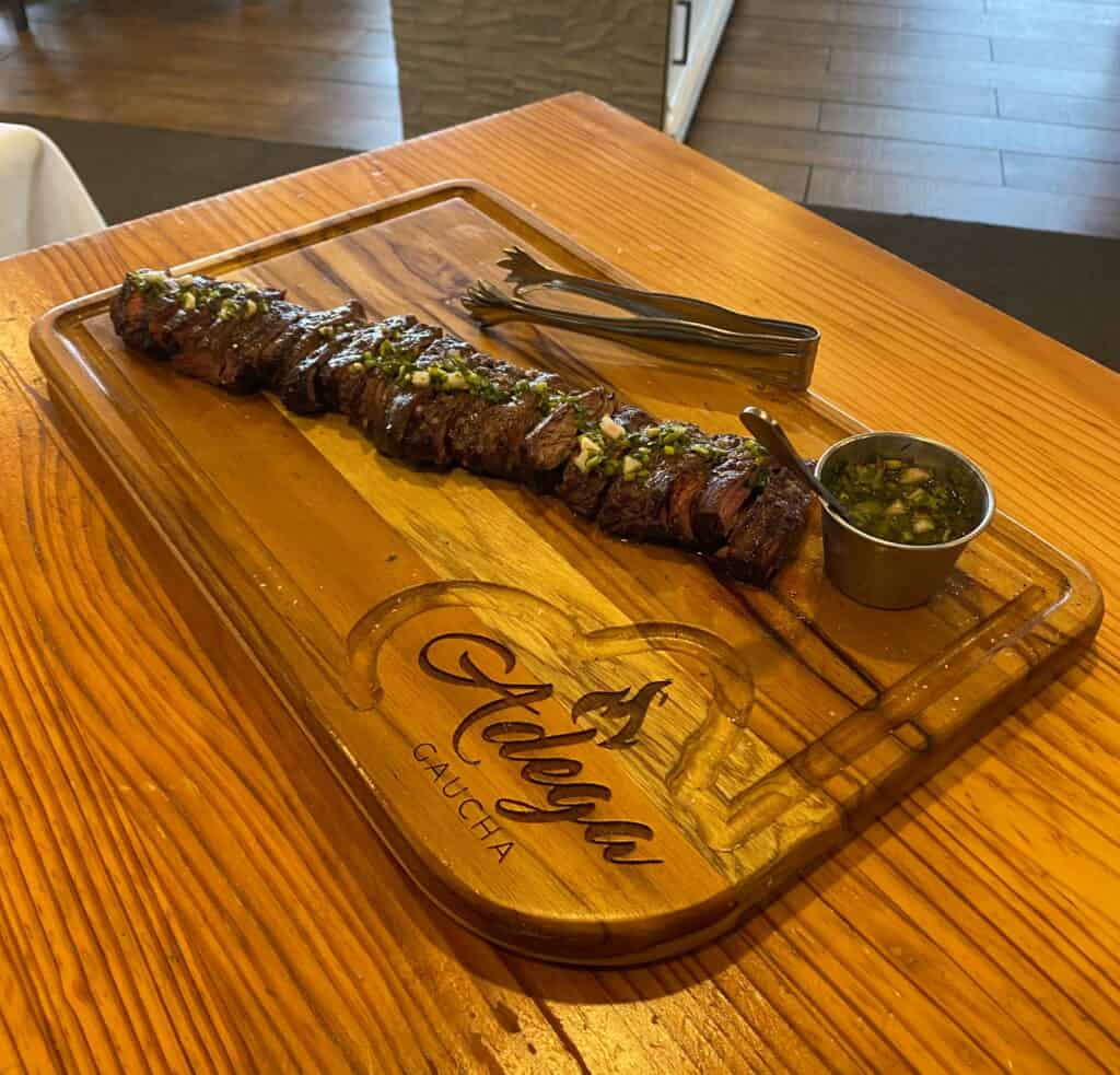 Sliced skirt steak with chimichurri served on a branded Adega Gaucha wooden cutting board next to a side of sauce and serving tongs.