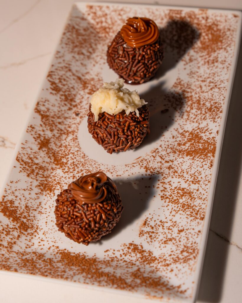 Three traditional Brazilian Brigadeiro chocolate truffles topped with chocolate, coconut, and dulce de leche on a white serving dish.